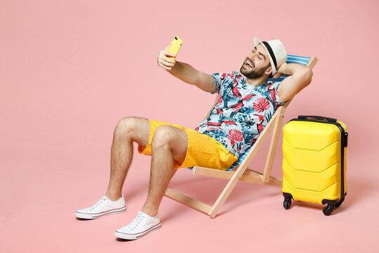 Full length cheerful young traveler tourist man in hat sit on deck chair doing selfie shot on mobile phone isolated on pink background studio. Passenger travel on weekend. Air flight journey concept.