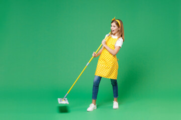 Full length of cheerful little kid girl housewife 12-13 in apron sing song in mop like microphone while doing housework isolated on green background children studio portrait. Housekeeping concept.