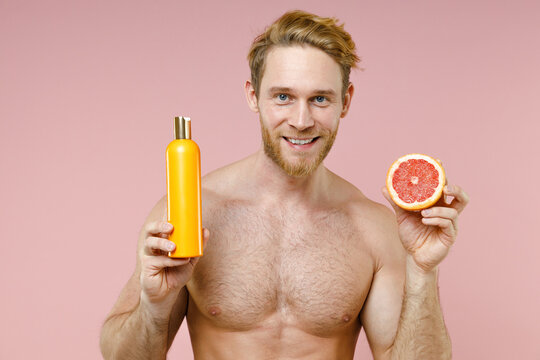 Funny bearded naked young man 20s perfect skin hold empty blank bottle of shampoo fresh grapefruit isolated on pastel pink background studio portrait. Skin care healthcare cosmetic procedures concept.