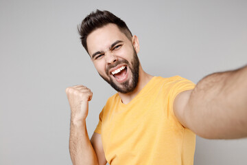 Close up young caucasian smiling bearded attractive man 20s in yellow basic t-shirt do selfie shot on mobile phone do winner gesture clench fist say yes isolated on grey background studio portrait.