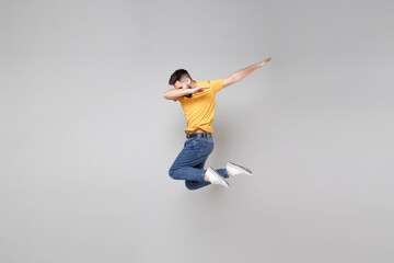 Fototapeta premium Full length of young bearded cool man 20s in yellow basic t-shirt jump high doing dab hip hop dance hands move gesture youth sign hiding covering face isolated on grey background studio portrait