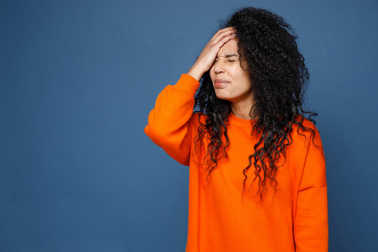 Confused young african american woman in casual basic bright orange sweatshirt standing put hand on face facepalm epic fail gesture keep eyes closed isolated on blue color background studio portrait.