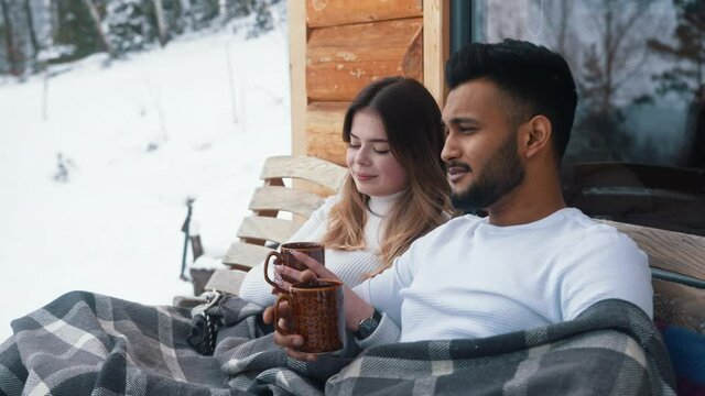 Happy young couple hugging covered with blanket on the outdoors bench. Drinking hot beverage and enjoying snow. High quality 4k footage
