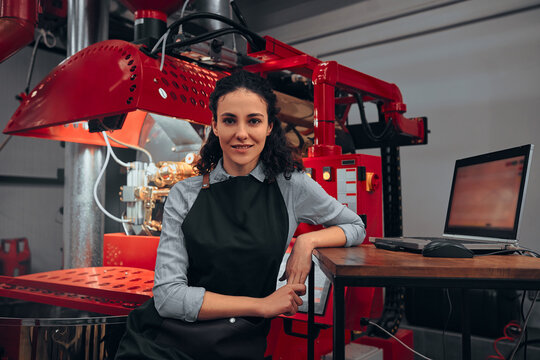 Woman controlling his laptop while operating a modern coffee roaster. Woman looking in camera. - Powered by Adobe