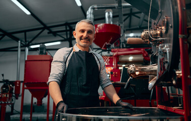 Portrait of smiling coffee roaster standing at roasting machine and looking at camera. Selective...