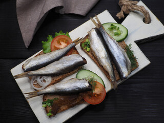Sandwiches with rye toast bread, smoked herring, vegetables and herbs on a white kitchen board, napkin, black wooden table, top view, flat layout. Gourmet seafood fish clupea harengus membras 