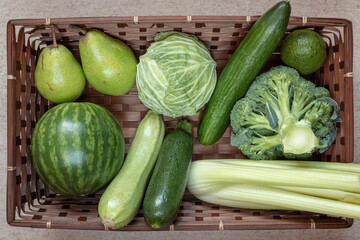 Flat lay composition of green fruits and vegetables including watermelon, pear, avocado, zucchini, cucumber, cabbage, broccoli, celery in brown rectangular basket