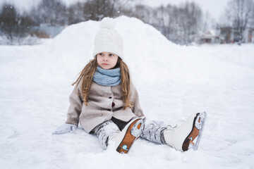 Fototapeta premium Beautiful little cute girl learn to skate on ice skating rink in park. Fall down and have fun. Stylish look, warm woolen coat, white hat, scarf, snood. Winter family activities, sport, games outdoors