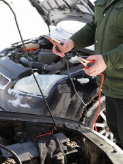 A Caucasian man mechanic stands with the starting wires near frozen snowy car engine, cold start with spare external battery accumulator and power wires with crocodile clips at Sunny winter day