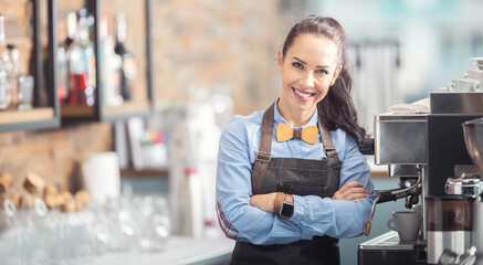Happy waitress in an apron and wooden bow tie stands confidently next to a coffee maker