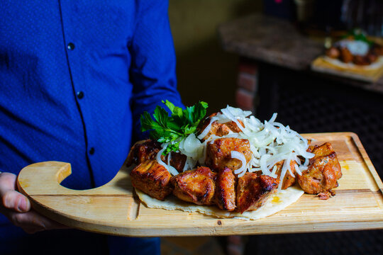 The Waiter Holds Fried Meat On A Wooden Board. Serving Barbecue With Herbs