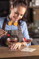 Smiling barmaid wearing wooden bow tie decorates manhattan cocktail drink with a cherry