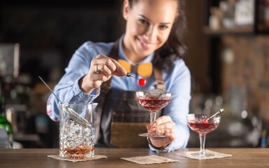 Smiling barmaid wearing wooden bow tie decorates manhattan cocktail drink with a cherry