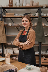 Vertical waist up portrait of young female artisan looking at camera while standing with arms crossed in workshop, hobby and small business concept - obrazy, fototapety, plakaty
