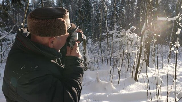 An elderly man takes photos of the winter sunny nature with a camera in the forest. There are trees and snowdrifts all around it.