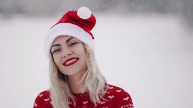 Portrait of cute woman in Christmas ugly sweater and Santa hat. Girl smiling, dancing, sending air kiss to camera.