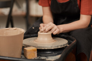 Warm-toned close up of female hands shaping clay on pottery wheel in workshop while enjoying arts and crafts, copy space