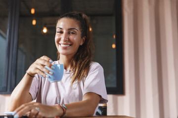 Beautiful smiling woman drinking tea on her home terrace, holding cup and looking happy at camera, sitting at coffee table in t-shirt