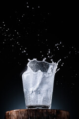 Pure mineral water in a glass, ice falls into the glass. Splashes of water rise above the glass, dark background with place for text, selective focus. Close-up, vertical frame