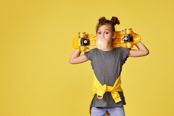 adorable little child girl with yellow skateboard blowing a big bubble gum over yellow background....