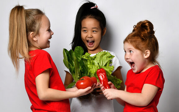 Children love to eat well. Happy little girls stand on a gray background with fresh vegetables and salad in their hands. The concept of healthy food for children and positive emotions.