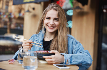 Eating out and food concept. Stylish blond girl eating outdoors in chinese restaurant, holding chopsticks and bowl, smiling at camera