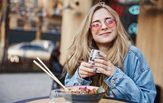 Happy woman eating outdoors chinese asian food, sitting on street table and drinking water, wearing sunglasses and smiling at camera