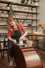 Vertical warm toned portrait of young female potter shaping clay on pottery whee while making ceramics in workshop and enjoying arts and crafts