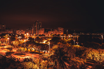 A long-exposure night cityscape in warm tones with a coastal Ipanema and Leblon districts of Rio de Janeiro, Brazil, a motion-blurred sky, highly illuminated beach, and plenty of residential houses