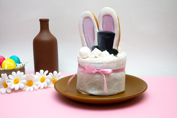 White cake with bunny ears and black hat in brown plate, three colorful eggs in nest, brown bottle on the white and pink background. Composition decorated with flowers