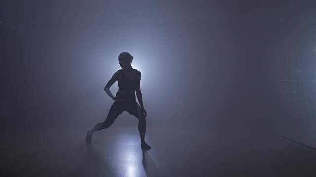 Silhouette of young woman dancing on dark stage with smoke and spotlight. Graceful dancer performing modern contemporary choreography. Full length shot in 4K, UHD