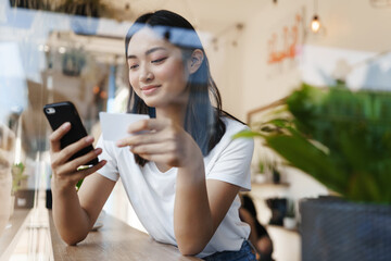 Stylish asian girl sitting in cafe near window, paying for online shopping with credit card, transfer money for coffee with smartphone, casually sitting in restaurant on summer day