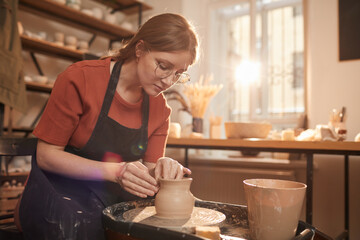 Warm toned portrait of young female artisan shaping clay on pottery wheel in sunlit workshop and...