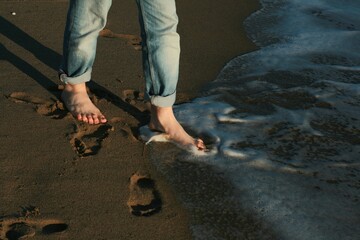 Close-up of the bare feet of a young man playing in the waves. He wears blue jeans rolled up. Vacation concept, enjoy, stroll on the beach. People enjoying walking on the sand.