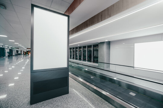 Wide-angle View Of A Vertical Empty Advertising Billboard Mockup Next To The Travelator Indoors Of A Modern Airport; A Template Of A Blank Information Poster In The Railroad Terminal, Moving Walkway