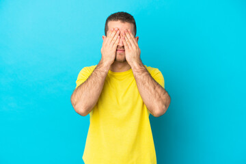 Young handsome caucasian man isolated on blue background covering eyes by hands
