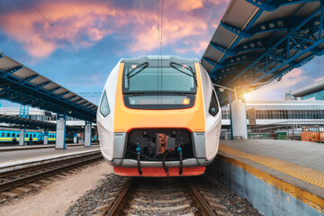 High speed train on the railway station at sunset. Industrial landscape with moving modern intercity passenger train on the railway platform, buildings. Railroad in Europe. Commercial transportation - obrazy, fototapety, plakaty