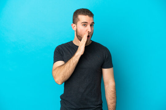 Young handsome caucasian man isolated on blue background whispering something with surprise gesture while looking to the side
