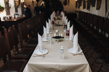 Table setting in the restaurant. White cotton tablecloth, napkins, glasses, plates and knives with forks, surrounded by wooden chairs. The concept of preparation for a celebration or party.