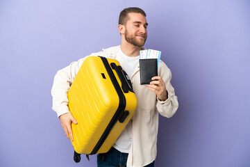 Young handsome caucasian man isolated on purple background in vacation with suitcase and passport