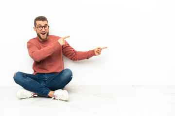 Young handsome man sitting on the floor surprised and pointing side