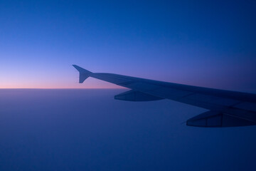 Fototapeta na wymiar The left side wing of a large white commercial airplane flying over the the Atlantic Ocean under the morning sun. The horizon is pink from the sunrise, the sky is blue and the ocean is dark blue. 