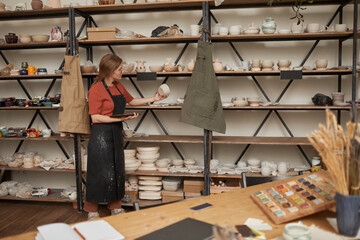 Background image of young female artisan organizing ceramics on shelves in pottery workshop, copy space
