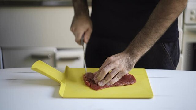A man cutting a piece of meat with a sharp knife over a white table. Indoors, kitchen.