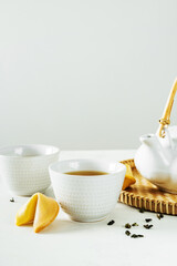 Asian tea concept, two white cups of tea, fortune cookies and teapot surrounded with green tea dry leaves with space for a text on light background