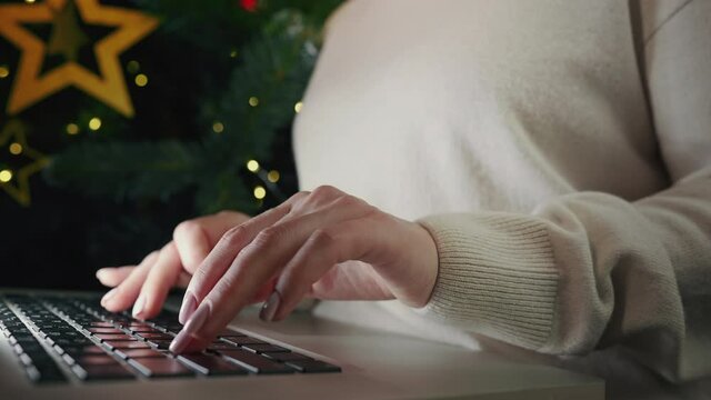 Business woman working on laptop, sitting near Christmas tree at home. Focus on hands typing on keyboard. Social distancing, creative workspace concept. Lady shopping online through website