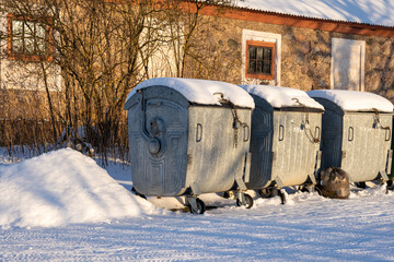 garbage containers stand next to each other in the winter sunlit where in the background you can see the masonry building