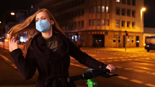 [4k] young woman with ffp2 mask on e-scooter is warm streetlight