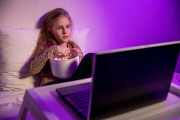 Little girl watching cartoon on laptop in bed in the evening and eating popcorn. She is surprised looking at the screen. Home entertainment. Evening twilight