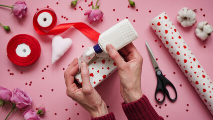 Step 8.Step-by-step instructions for wrapping gifts for Valentine's Day. Woman wraps a gift with wrapping white paper in heart and seals the edges with glue on a pink background top view, flat lay